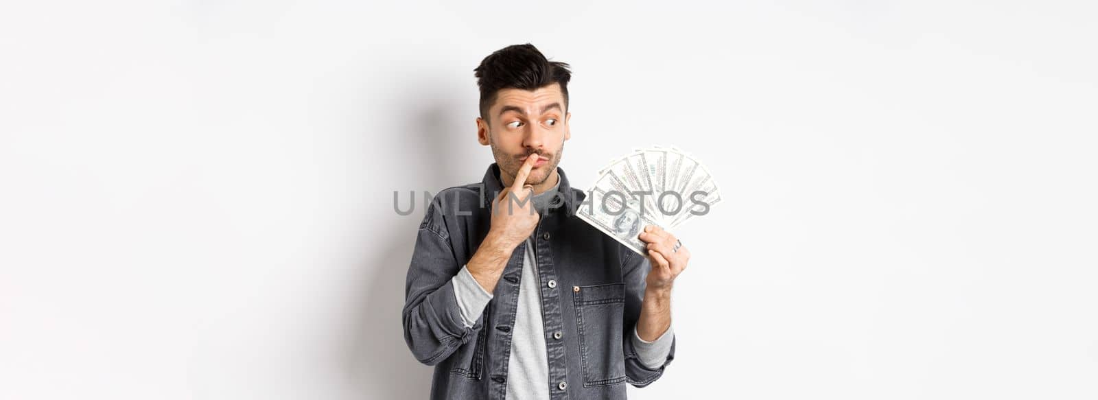 Man thinking of shopping while looking at dollar bills, holding money and staring pensive, standing on white background.