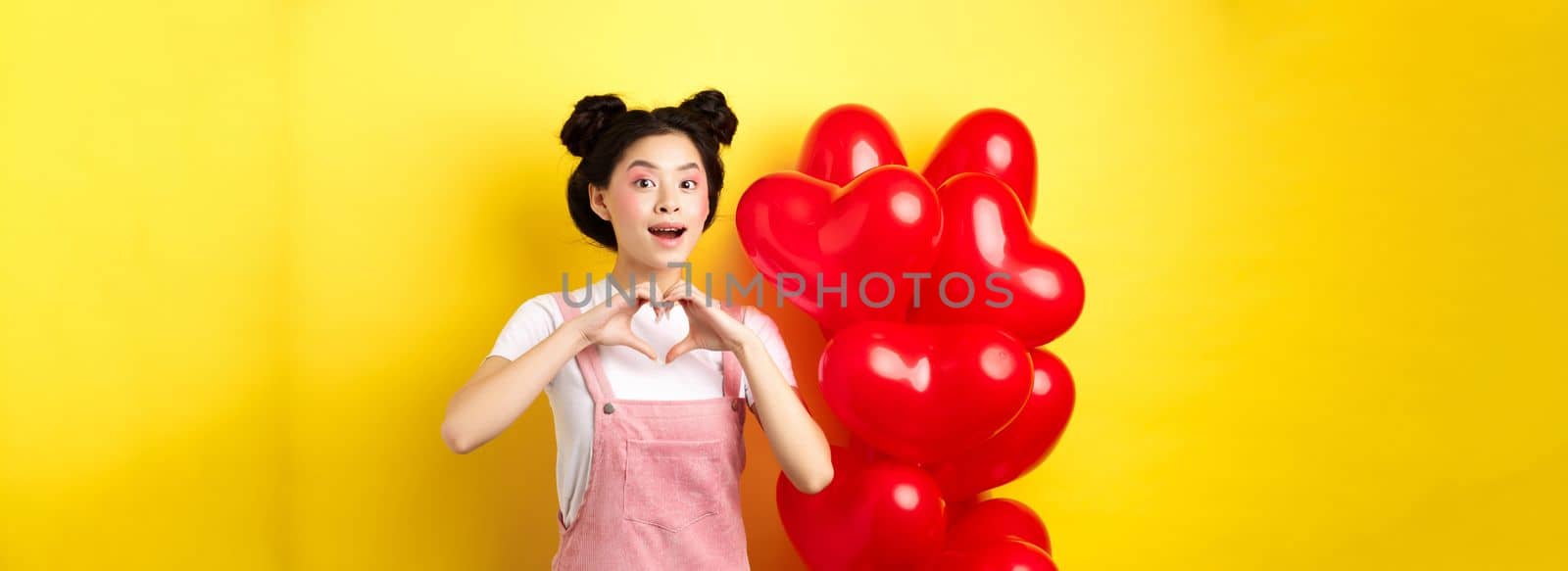 Happy asian girl showing heart sign near romantic balloons, waiting for true love on Valentines day, looking hopeful at camera, yellow background by Benzoix