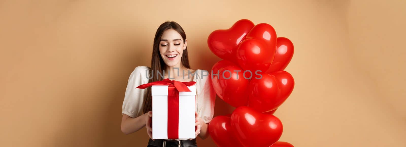 Happy woman open Valentines day gift, smiling excited and looking at present box, standing near red heart balloons on beige background.