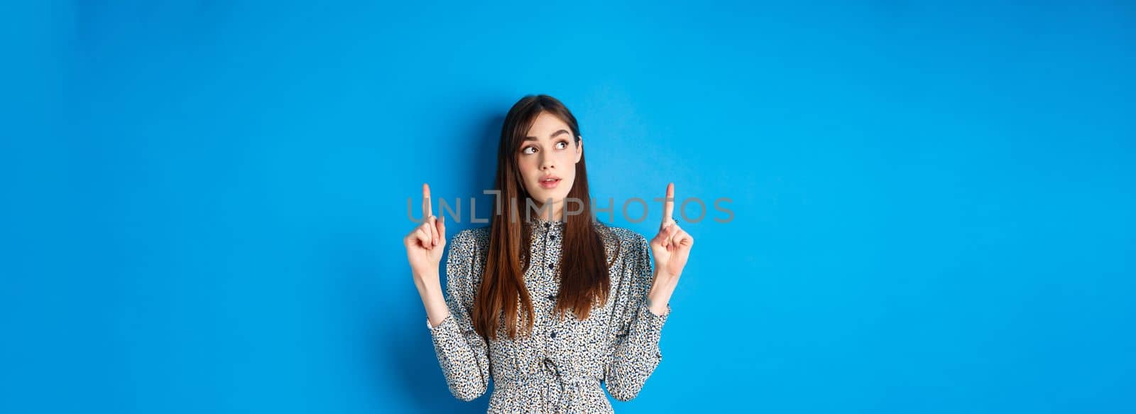 Pretty thoughtful woman pointing fingers up and thinking, standing pensive in dress on blue background.