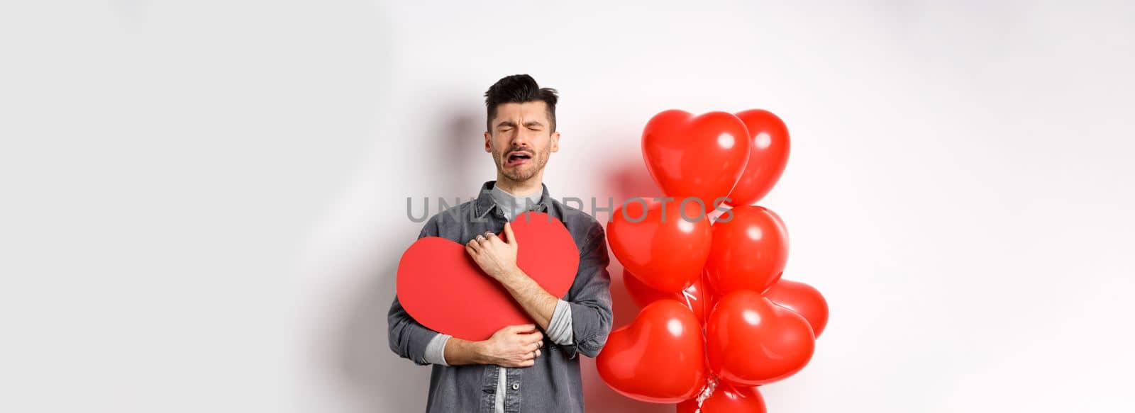 Crying man standing single and lonely on Valentines day, hugging heart cutout and sobbing miserable, being heartbroken and rejected by lover, white background by Benzoix