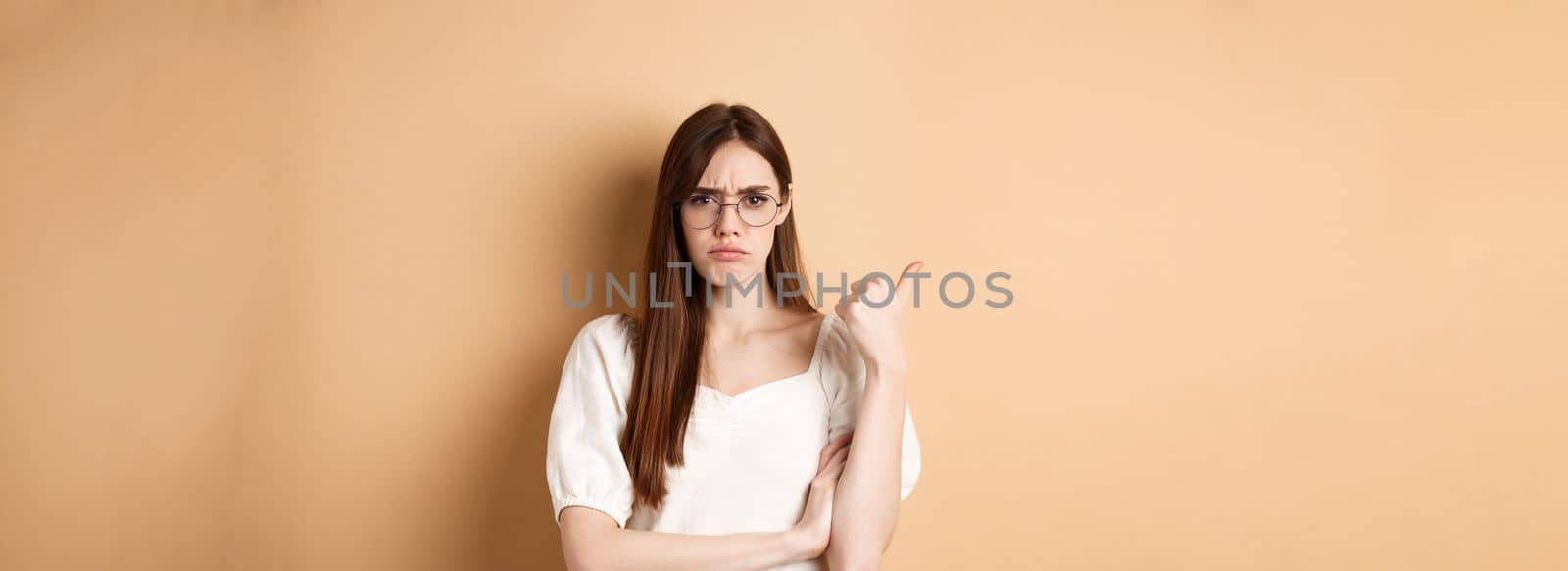 Disappointed girl in glasses frowning, pointing aside at bad product, disapprove and dislike something, standing on beige background.