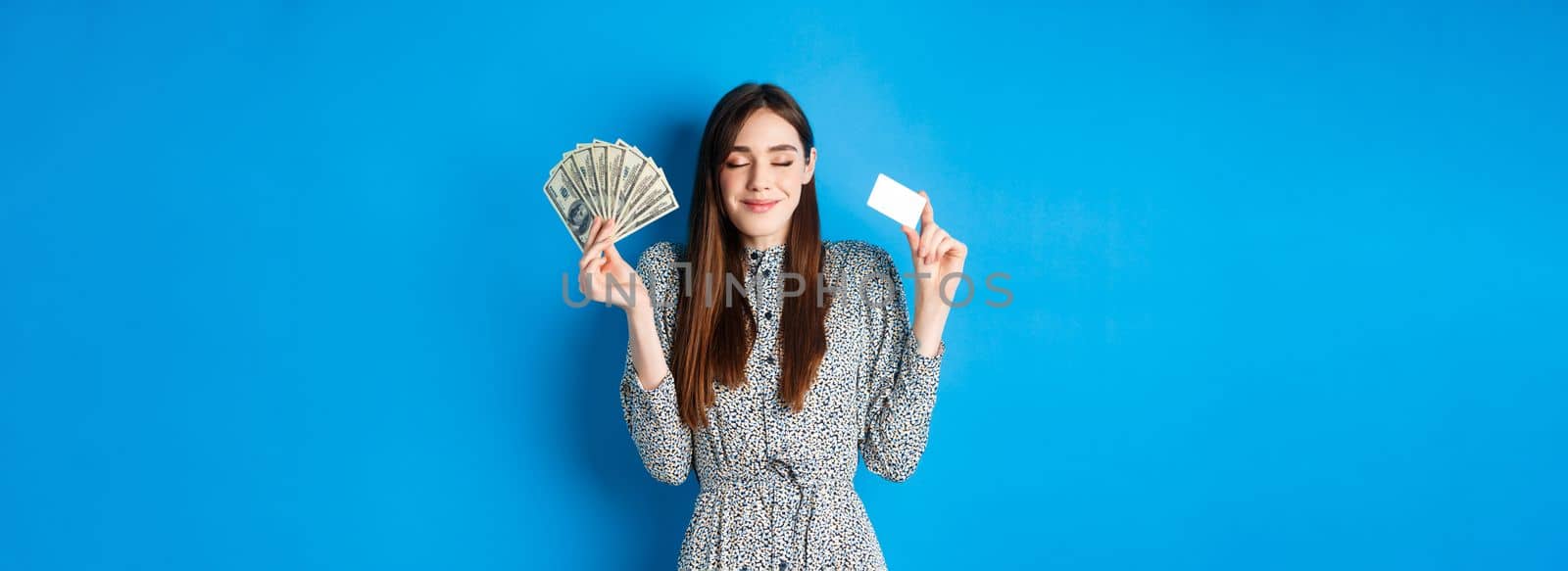 Shopping. Happy smiling woman looking satisfied with eyes closed, showing money dollar bills and plastic credit card, standing dreamy against blue background.