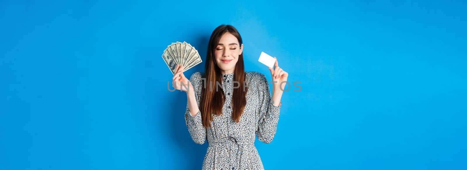 Shopping. Happy smiling woman looking satisfied with eyes closed, showing money dollar bills and plastic credit card, standing dreamy against blue background by Benzoix