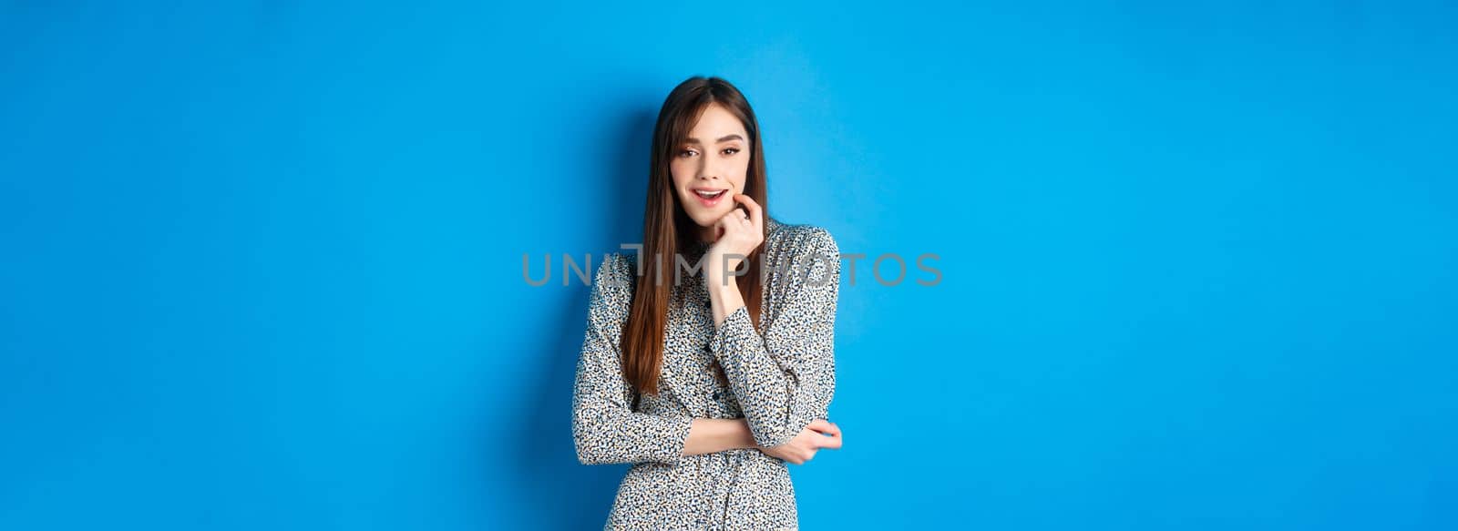 Coquettish girl in trendy long dress looking excited at camera, standing on blue background.