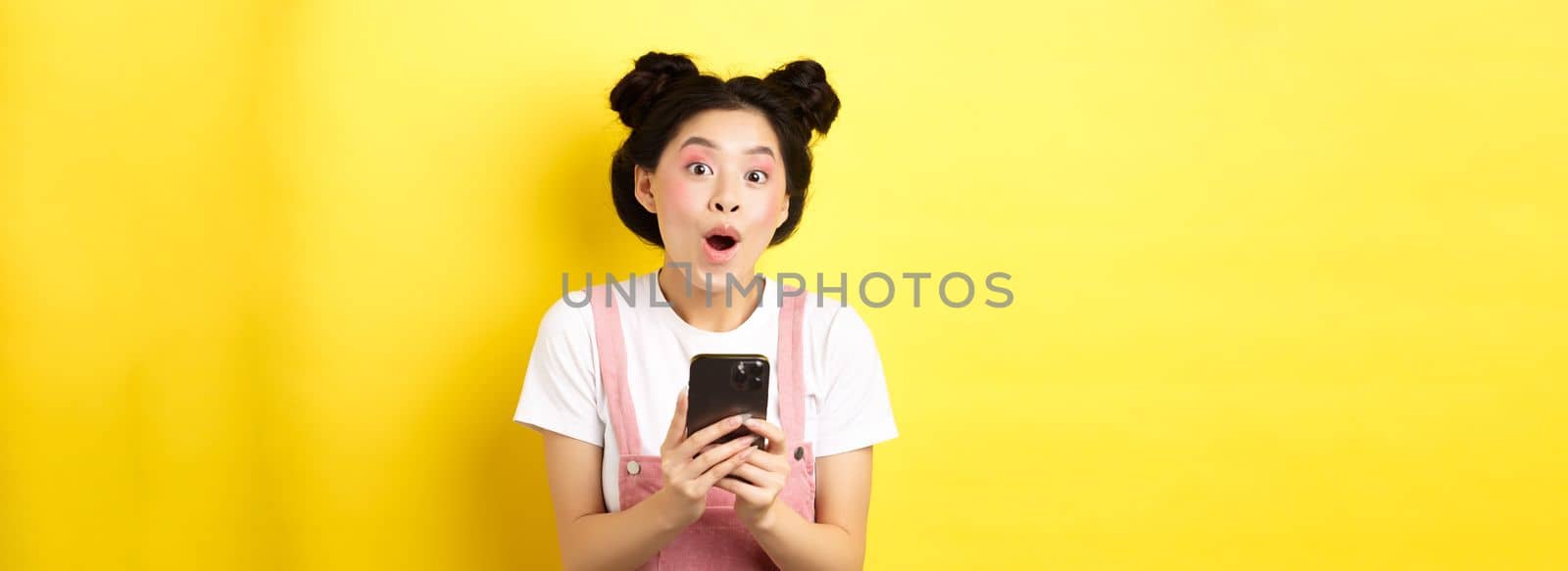 Excited girl with glam makeup and summer clothes, gasping amazed, checking out internet offer on mobile phone, standing on yellow background by Benzoix