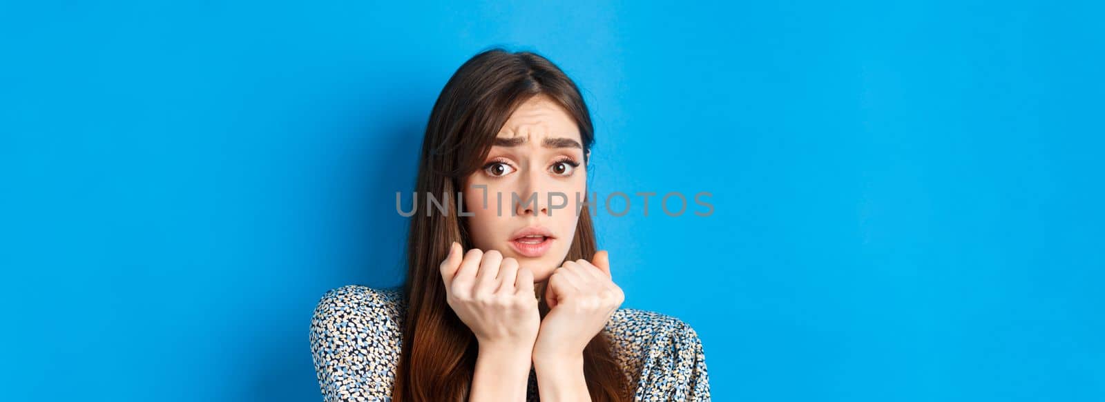 Close-up of scared timid girl looking afraid, shaking from fear, press hands to body and staring frightened, standing on blue background.