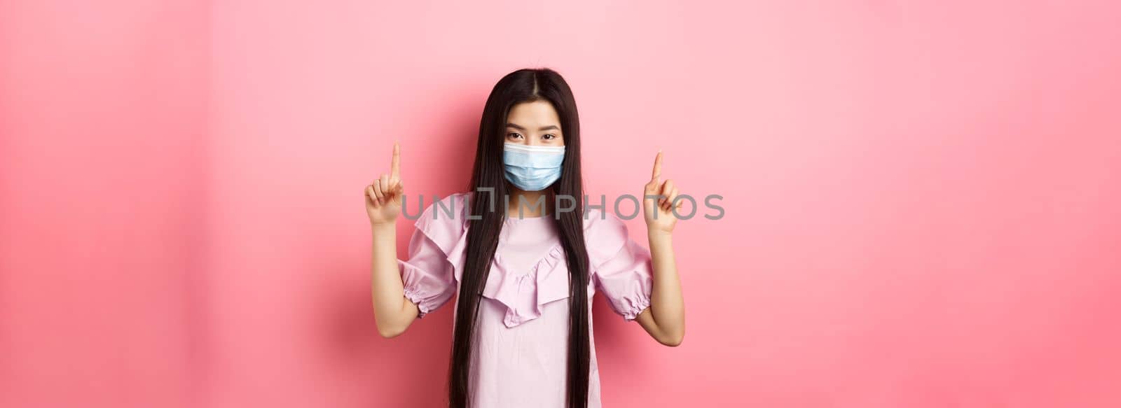 Coronavirus, quarantine and lifestyle concept. Beautiful asian woman in sterile medical mask pointing fingers up, smiling with eyes and look confident, standing against pink background.