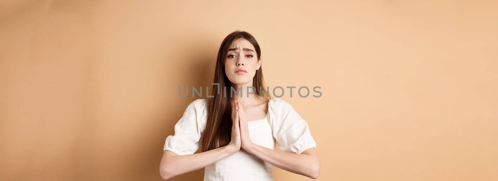 Please help. Desperate young woman begging you, holding hands in pleading gesture and looking sad at camera, asking for favour, beige background by Benzoix