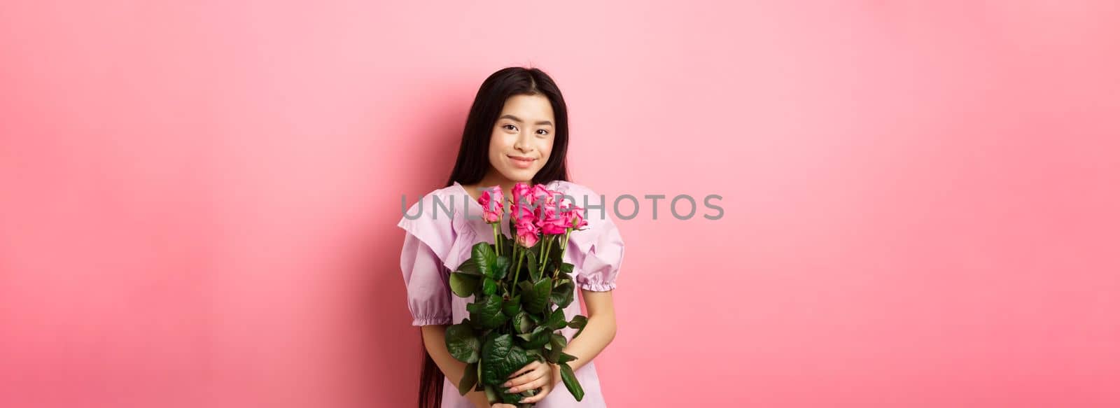 Romantic tender asian girl holding bouquet of roses, smiling cute at camera, having valentines date with lover, wearing dress, pink background.