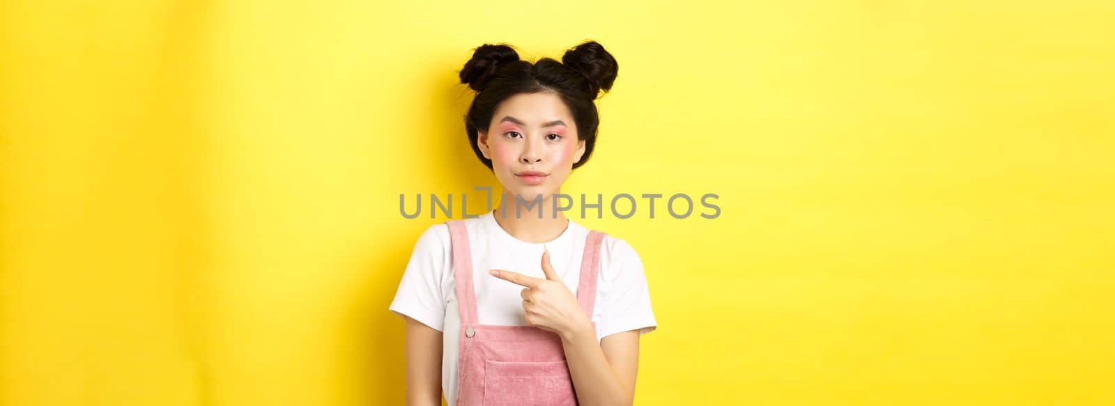 Stylish asian teen girl with makeup and summer clothes, pointing finger right and look serious, standing against yellow background.