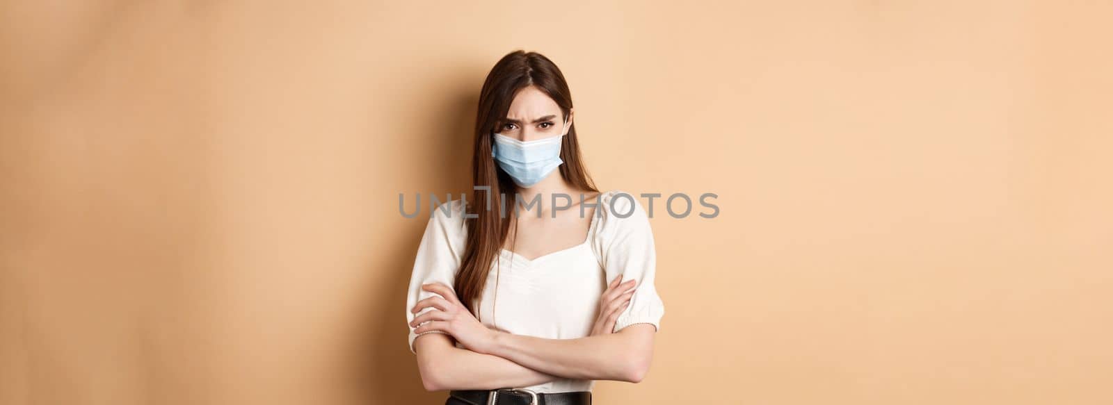 Covid-19 and lifestyle concept. Angry woman look with despise, wearing medical mask, cross arms on chest and stare with condemn, standing on beige background.