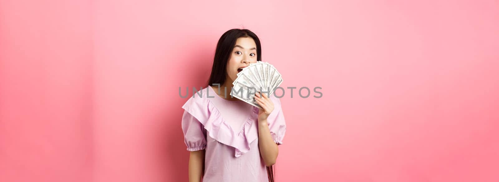 Cute asian woman showing dollar bills and smiling amazed. Rich woman waiving with money, standing against pink background by Benzoix