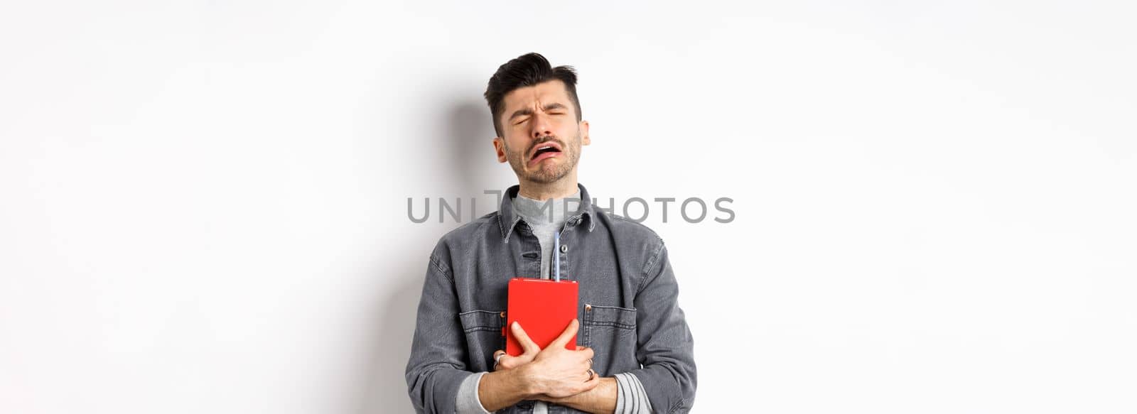Sad cryig man holding red diary and sobbing, miserable guy carry journal with him, standing against white background by Benzoix