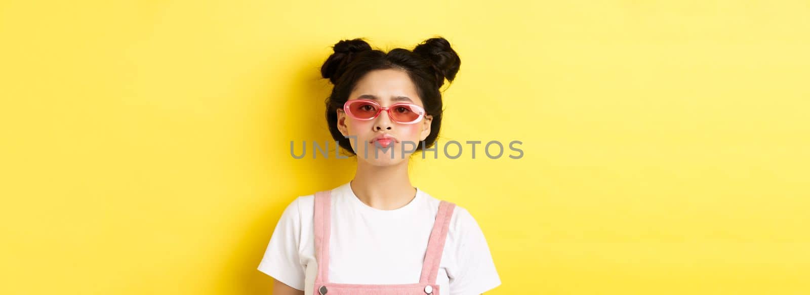 Summer and fashion concept. Bored asian teen girl in sunglasses pouting, standing moody on yellow background.