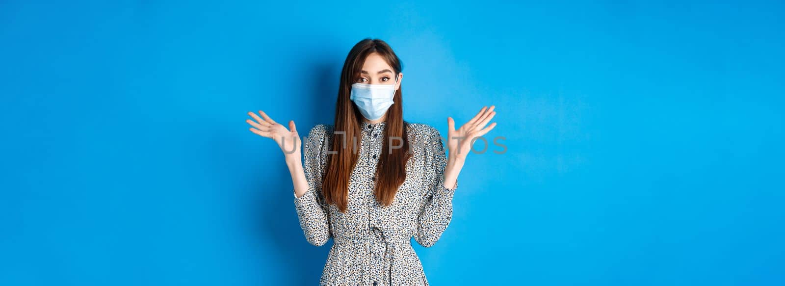 Healthy people and covid-19 pandemic concept. Surprised girl in medical mask raising hands up excited, hear good news, smiling with eyes, standing blue background.