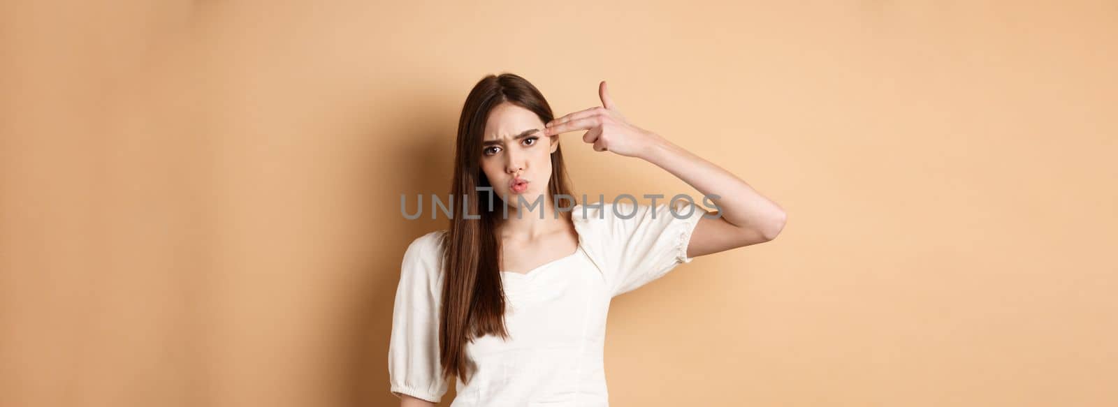 Just kill me. Annoyed and distressed young woman making finger gun sign on ver head and frowning bothered, standing pissed-off on beige background.
