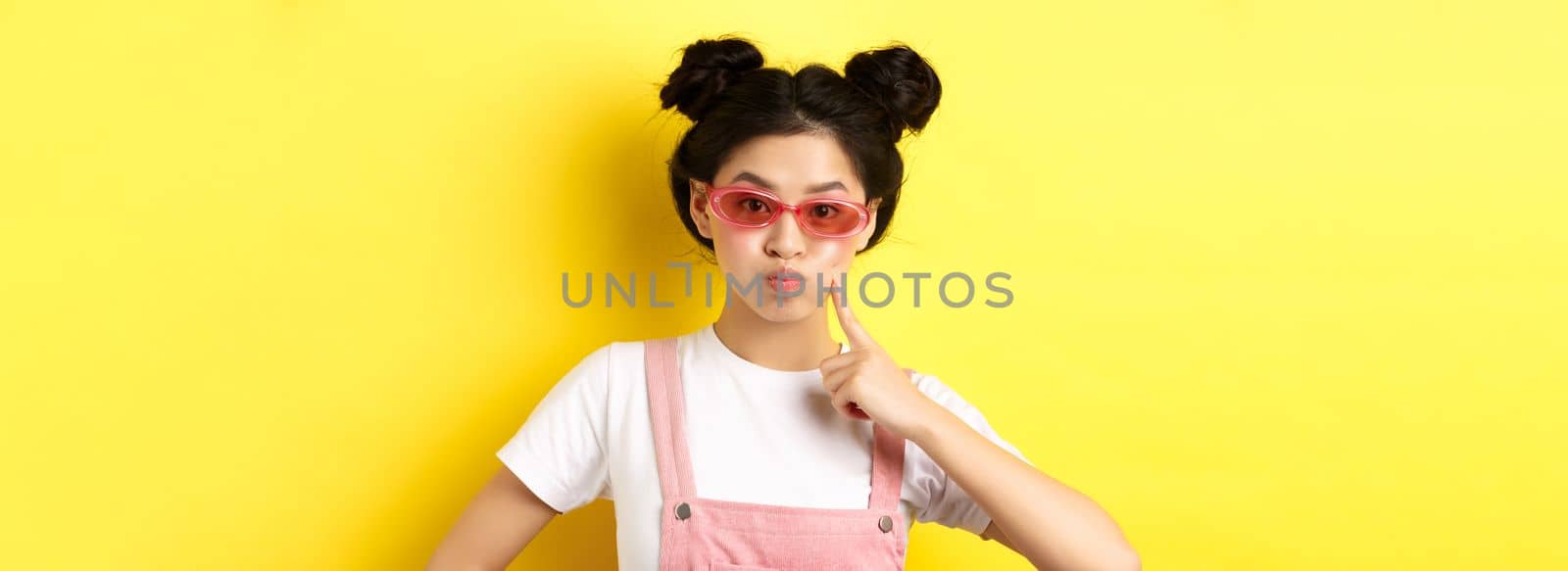 Stylish glam girl with sunglasses, pouting and poking cheek, standing silly on yellow background by Benzoix