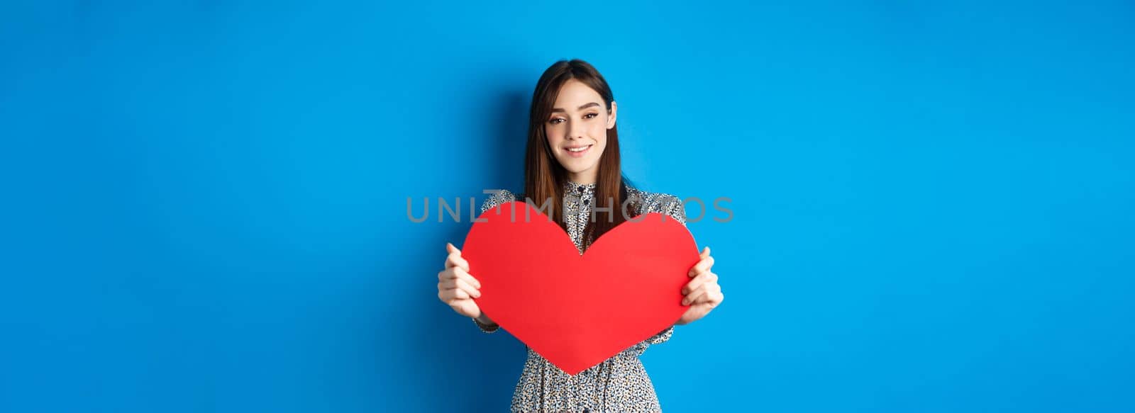 Valentines day and relationship concept. Beautiful romantic girl say I love you, extending hand with big red heart, standing on blue background.