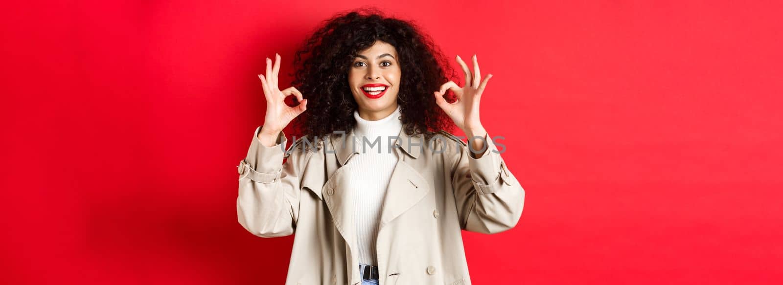 Stylish caucasian woman in trench coat showing okay gesture and smiling satisfied, recommending company, standing on red background.