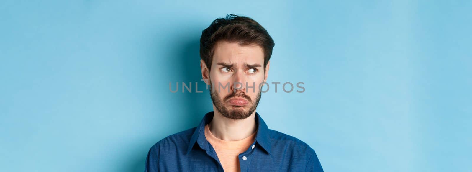 Miserable and disappointed guy acting childish, sulking and looking left at empty space with offended face, standing on blue background.