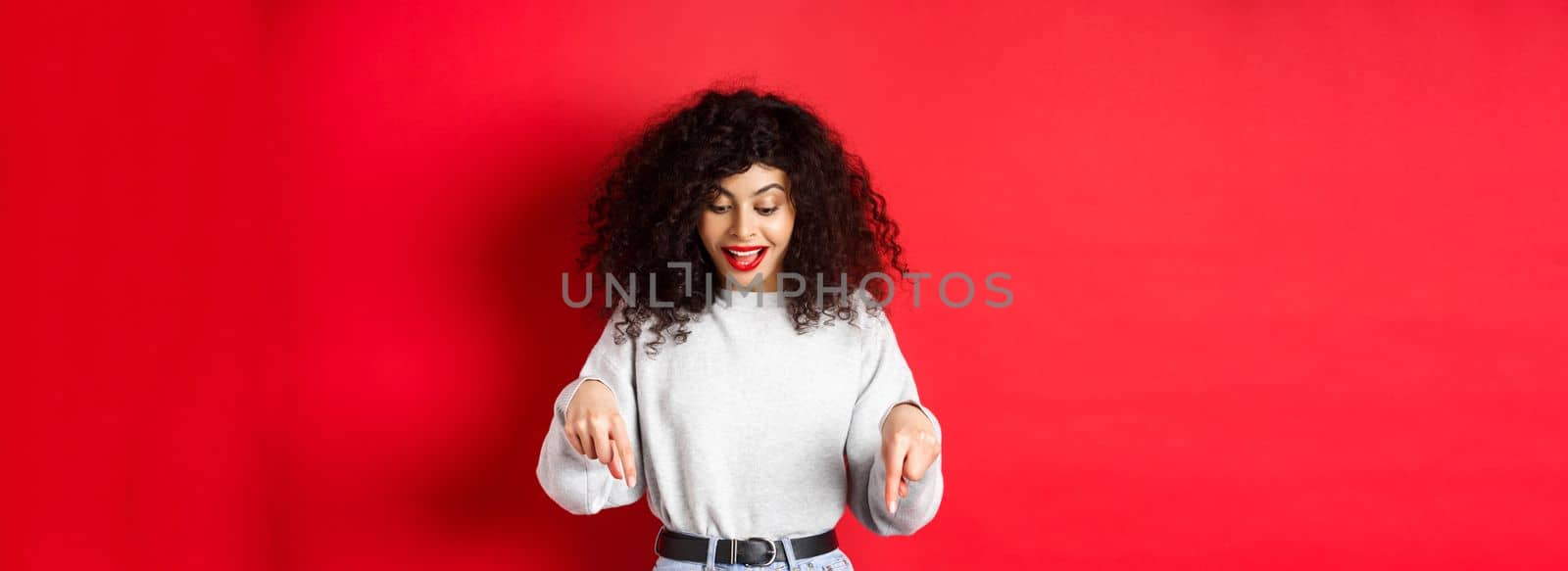 Dreamy beautiful woman with curly hair, pointing and looking down excited, checking out promo, standing against red background.