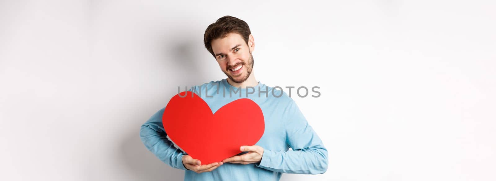 Charming lover man holding Valentines day red heart and smiling, I love you gesture, standing over white background by Benzoix