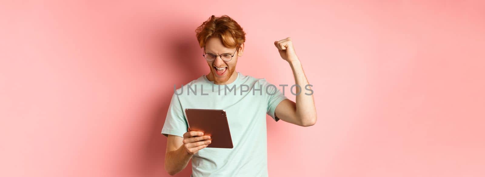 Image of happy redhead man triumphing, winning online with digital tablet and rejoicing, standing over pink background by Benzoix