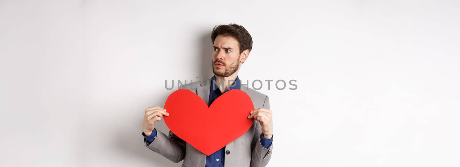Confused man in suit holding big red heart and looking right, searching for love on Valentines day, standing over white background.
