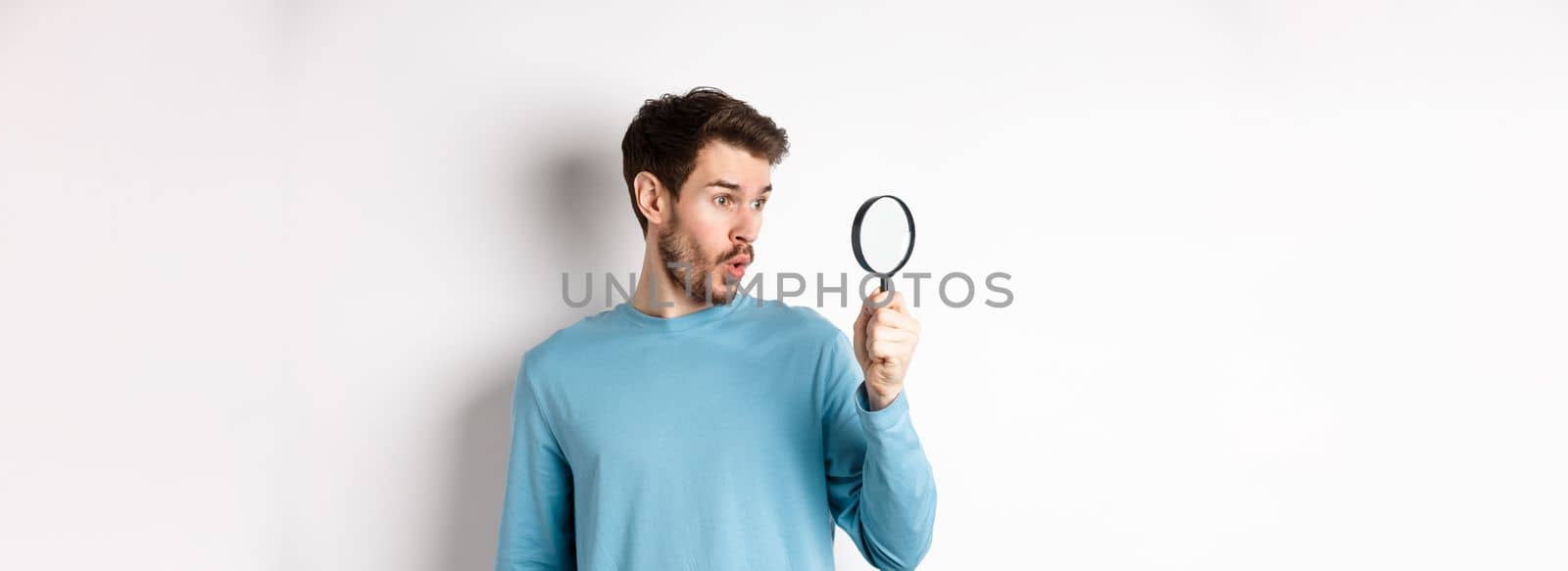 Man got surprised while looking through magnifying glass, saying wow at awesome product, standing over white background.