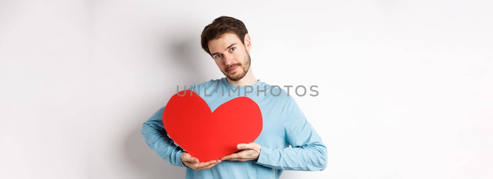 Romantic boyfriend making Valentines day surprise, holding big red heart cutout on chest and smiling with love, looking tender at camera, standing over white background by Benzoix