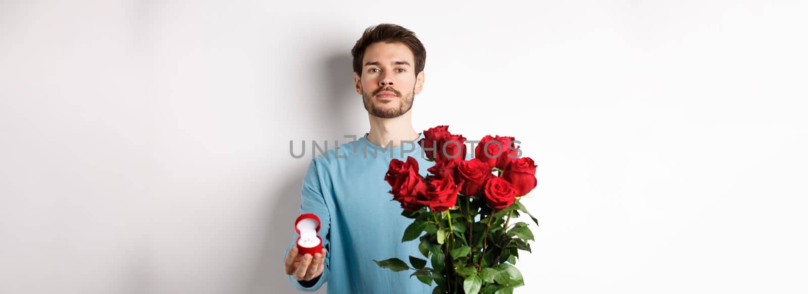 Valentines and relationship. Romantic man boyfriend holding red roses and showing engagement ring, making a proposal on lovers day, standing over white background by Benzoix