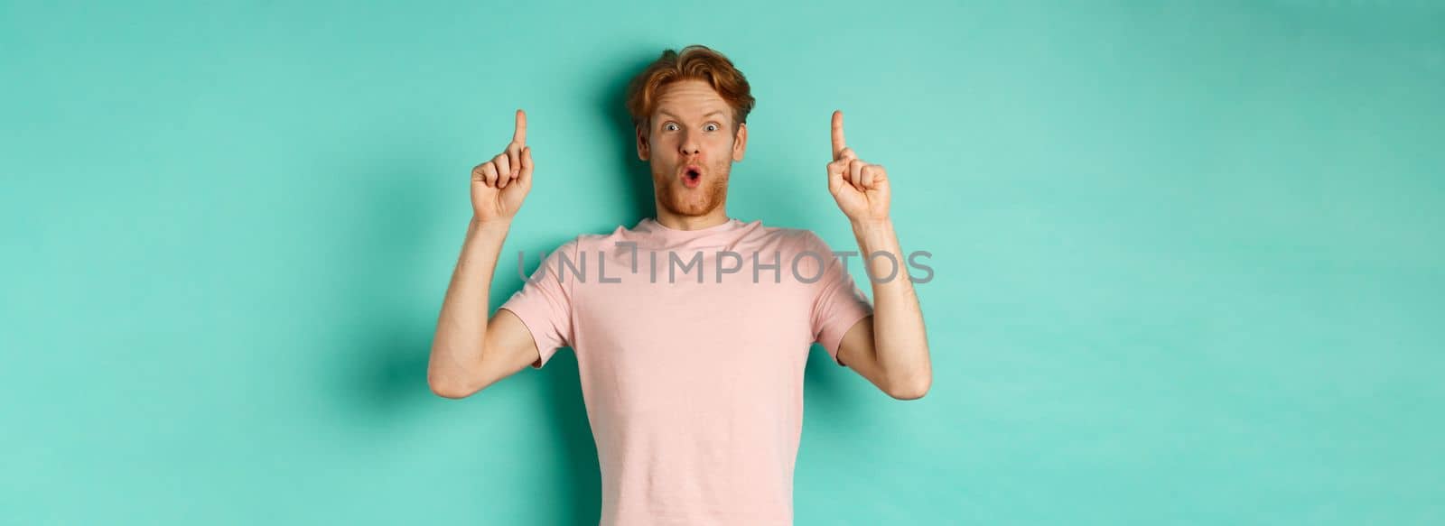 Impressed handsome guy with red hair pointing fingers up, demonstrate promo offer, standing in t-shirt over mint background.