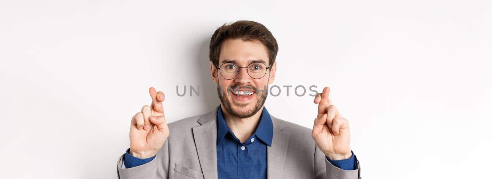 Close-up of hopeful male entrepreneur in glasses and suit cross fingers for good luck, smiling and making wish, praying for good results, white background.