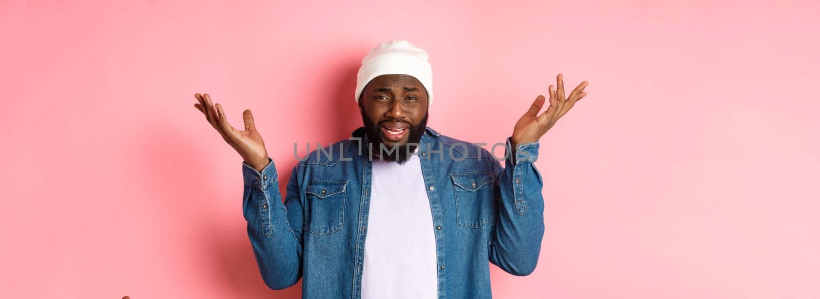 Confused and bothered Black man complaining, shrugging and raising hands up, staring with disappointment, standing over pink background.