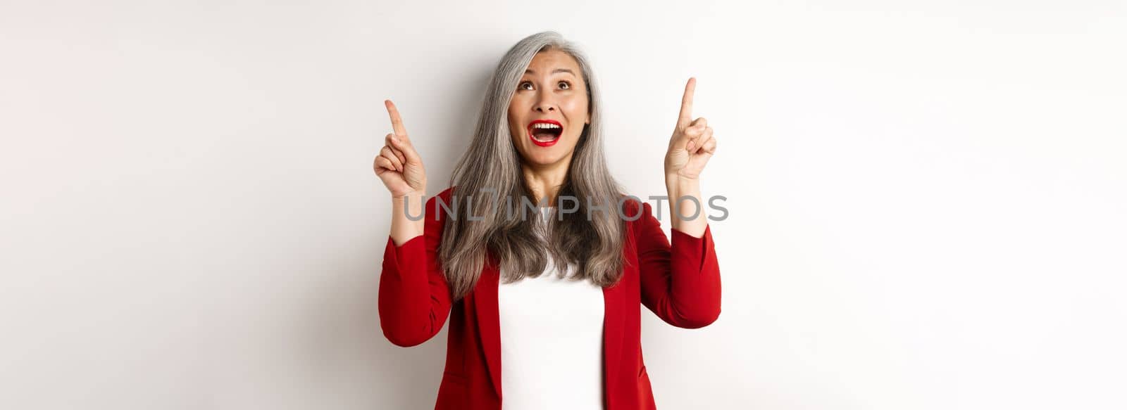 Happy asian lady in red blazer and makeup, looking and pointing fingers up, showing special offer, standing over white background.