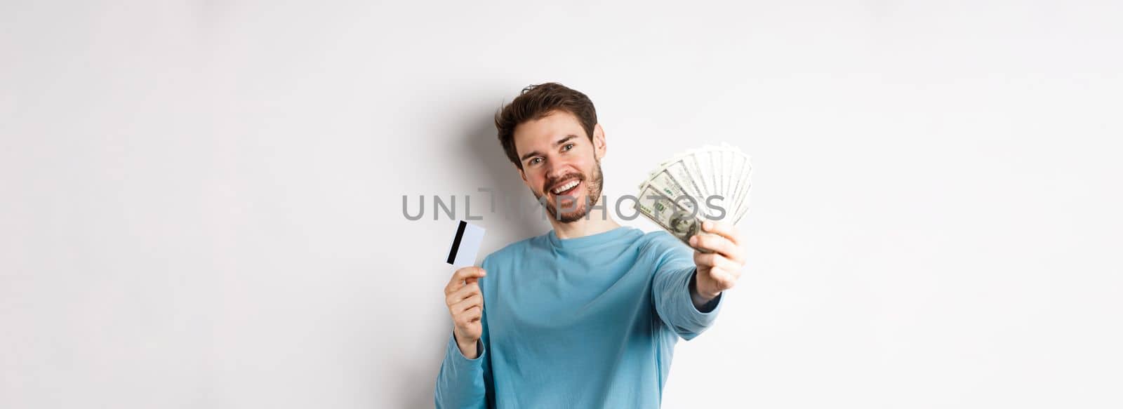 Handsome young man in blue shirt giving you money, holding plastic credit card, standing over white background.