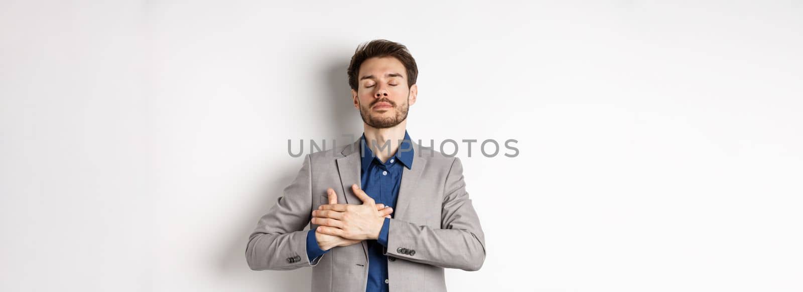 Young man in suit close eyes, holding hands on heart and remember happy memories, feeling nostalgic, standing against white background by Benzoix