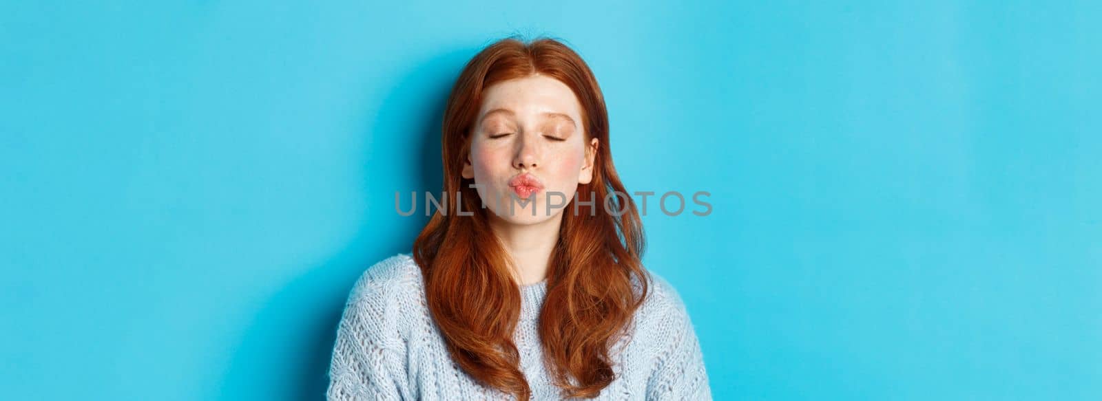 Close-up of young woman with red wavy hair standing with eyes closed and puckered lips, waiting for kiss against blue background by Benzoix
