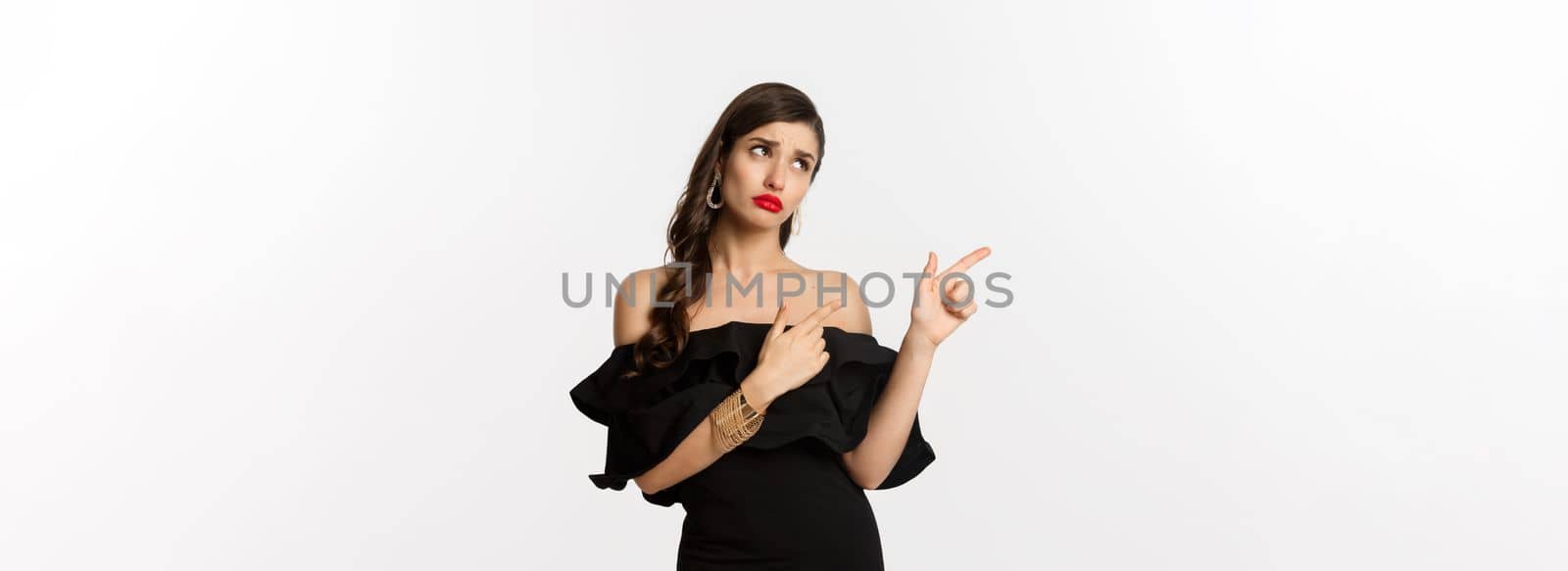 Fashion and beauty. Jealous glamour woman in black dress looking and pointing fingers left, sulking disappointed, white background.