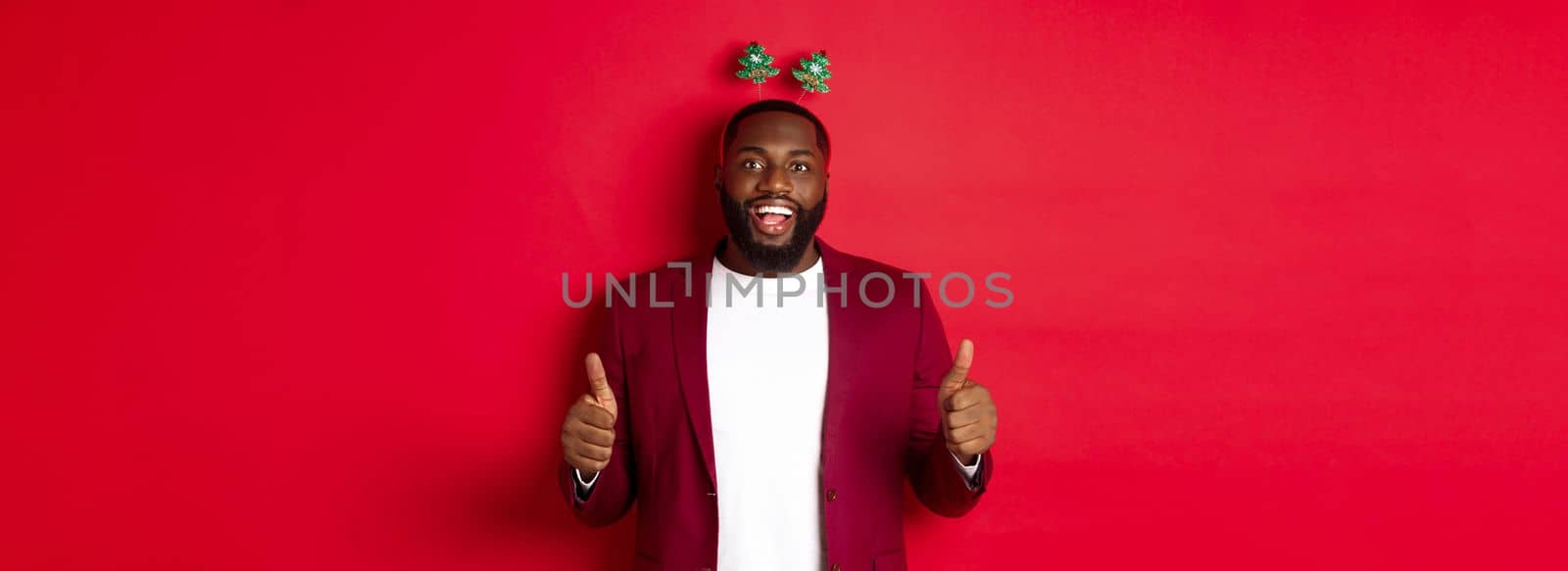 Merry Christmas. Happy african american man celebarting New Year, wearing funny party headband and showing thumb up, like and praise something, standing over red backgrond.