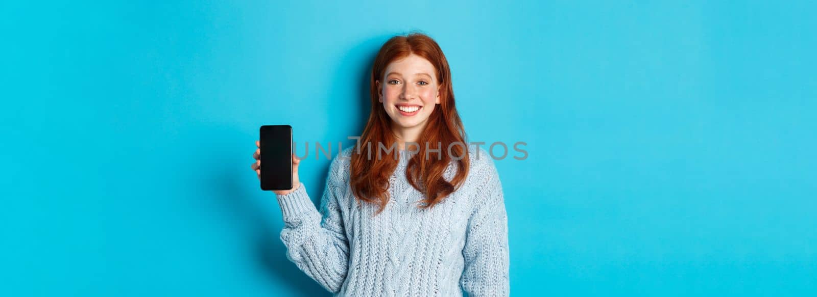 Smiling female model with red hair showing smartphone screen, holding phone and demonstrating application, standing over blue background.