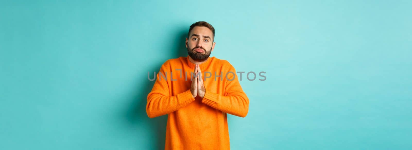 Sulking man begging for help, asking for favour, holding hands in pray and pouting, standing over turquoise background.