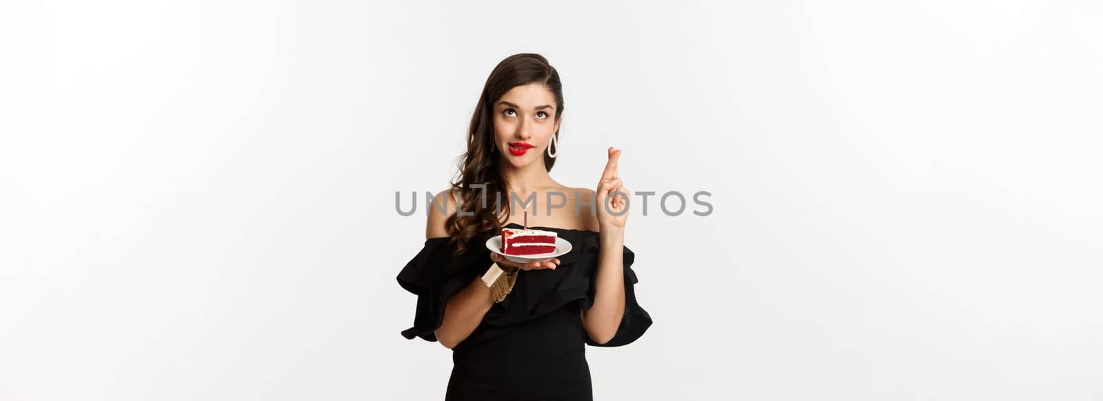 Celebration and party concept. Hopeful and dreamy woman making wish on birthday cake, cross fingers and smiling happy, standing over white background.