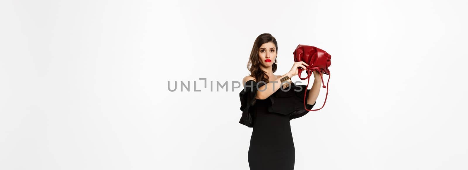 Beauty and fashion concept. Full length of sad young woman in black dress and high heels showing empty purse, sulking disappointed, standing over white background.