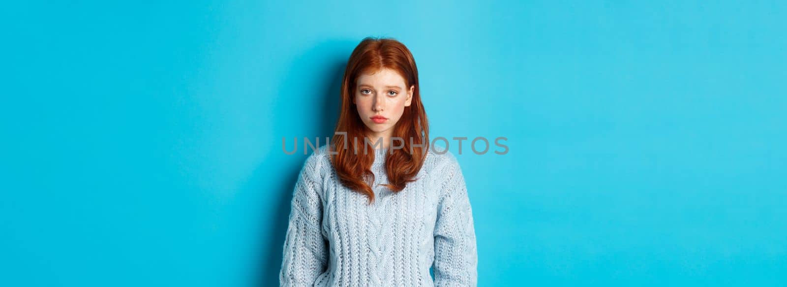 Sad and gloomy redhead teenage girl staring at camera uneasy, feeling bad, standing against blue backgorund in sweater.