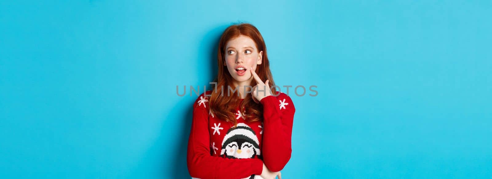 Winter holidays and Christmas Eve concept. Pretty redhead girl in xmas sweater, touching cheek thoughtful and smiling, making choice, looking at upper right corner and thinking, blue background.
