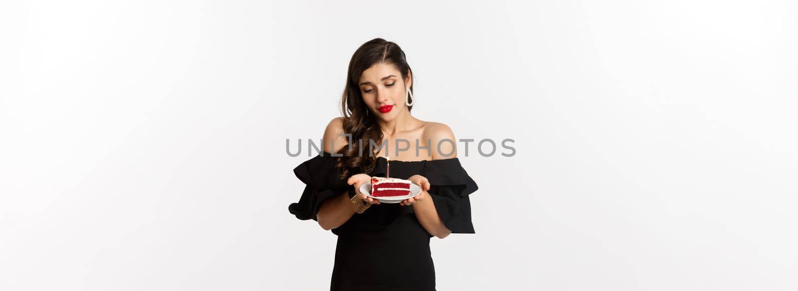 Party and celebration. Cute young woman with red lips, wearing black dress, holding birthday cake with candle and looking dreamy, making wish, white background by Benzoix