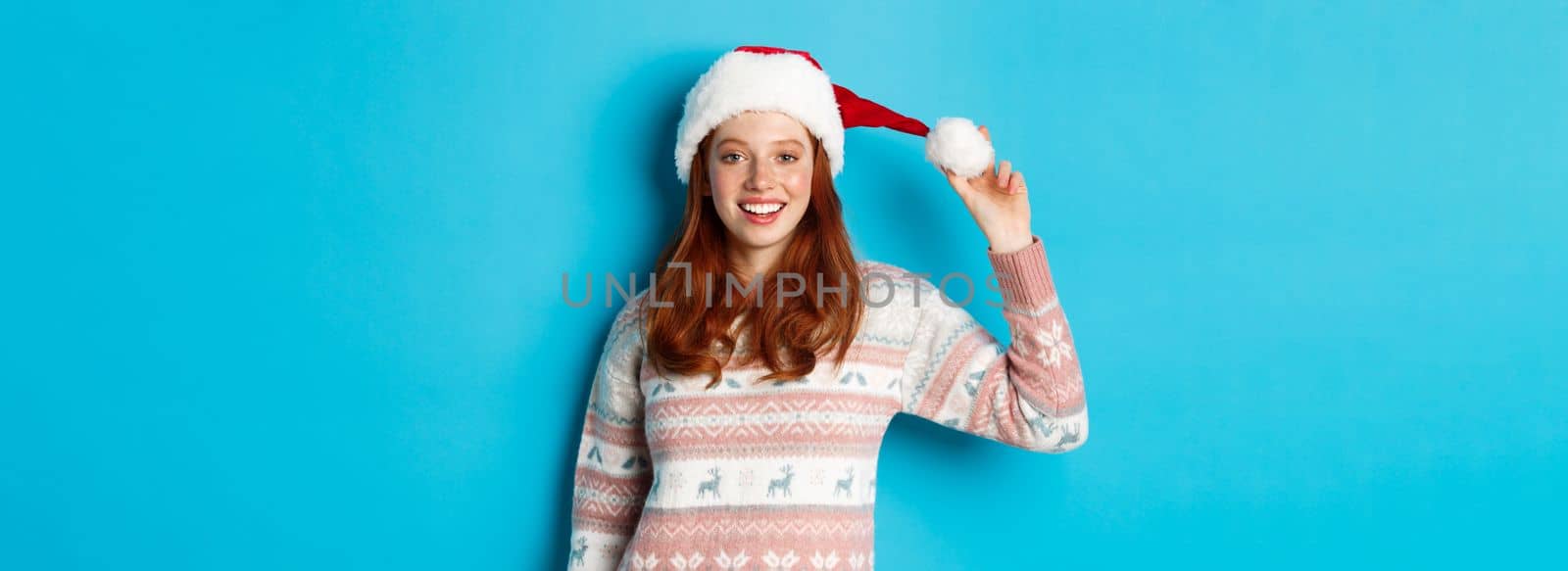 Winter and Christmas Eve concept. Cute redhead girl in Santa hat smiling at camera, standing in sweater against blue backgroudn.