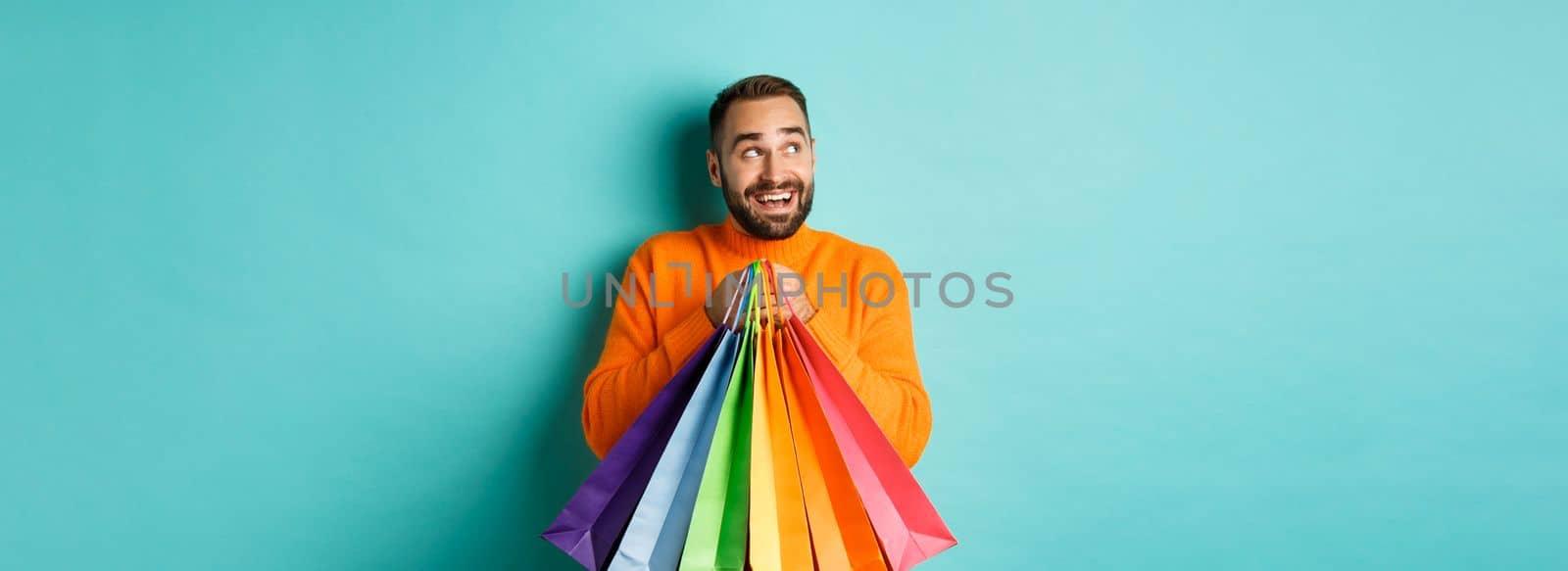 Happy caucasian man in orange sweater, looking left and imaging, holding shopping bags and smiling, standing over turquoise background.